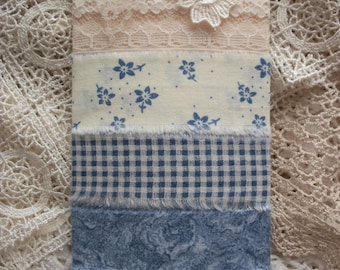 May include: A rectangular tag wrapped with five horizontal fabric strips featuring cream lace with a floral applique, a cream fabric with blue flowers, a blue and white gingham check, a blue mottled pattern, and a dark blue fabric with tiny dots.