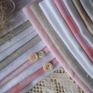 May include: An assortment of fabric swatches in shades of white, pink, and beige, arranged in diagonal lines. Three small, round, off-white buttons are placed on top of the fabric. The fabrics have various textures, including linen and woven patterns. A piece of lace is visible in the background.