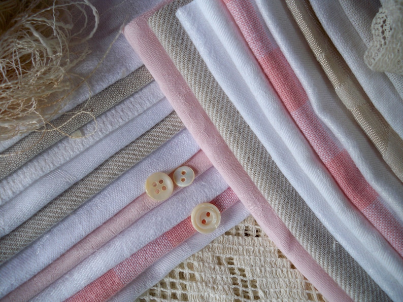 May include: An assortment of fabric swatches in shades of white, pink, and beige, arranged in diagonal lines. Three small, round, off-white buttons are placed on top of the fabric. The fabrics have various textures, including linen and woven patterns. A piece of lace is visible in the background.