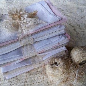 May include: A stack of folded white, pink, and gray fabric squares tied with lace ribbon and adorned with a flower and buttons. Two balls of beige thread and loose thread are next to the stack. The items are on a lace surface.