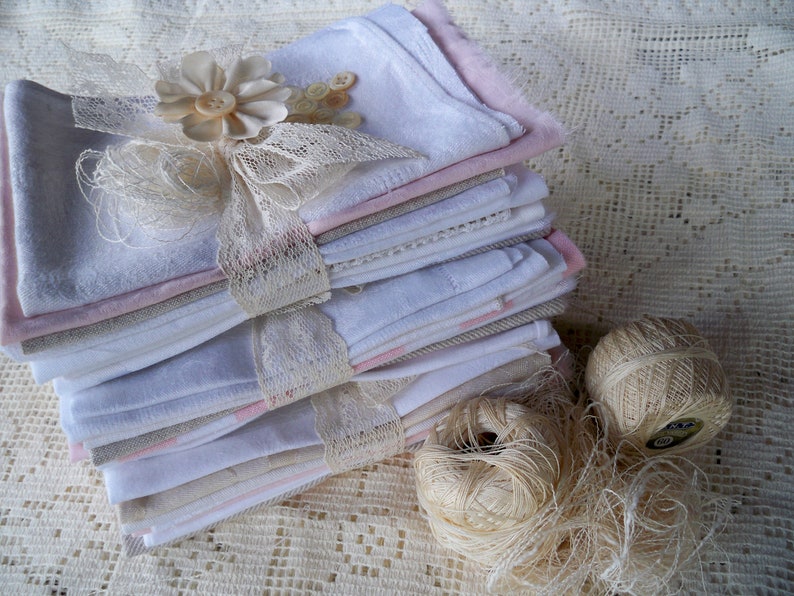 May include: A stack of folded white, pink, and gray fabric squares tied with lace ribbon and adorned with a flower and buttons. Two balls of beige thread and loose thread are next to the stack. The items are on a lace surface.