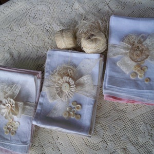 May include: Three stacks of white linen napkins, each adorned with lace, ribbon, and buttons. The napkins are decorated with floral accents and tied with lace. Two spools of twine are also visible, suggesting a craft or home decor theme.