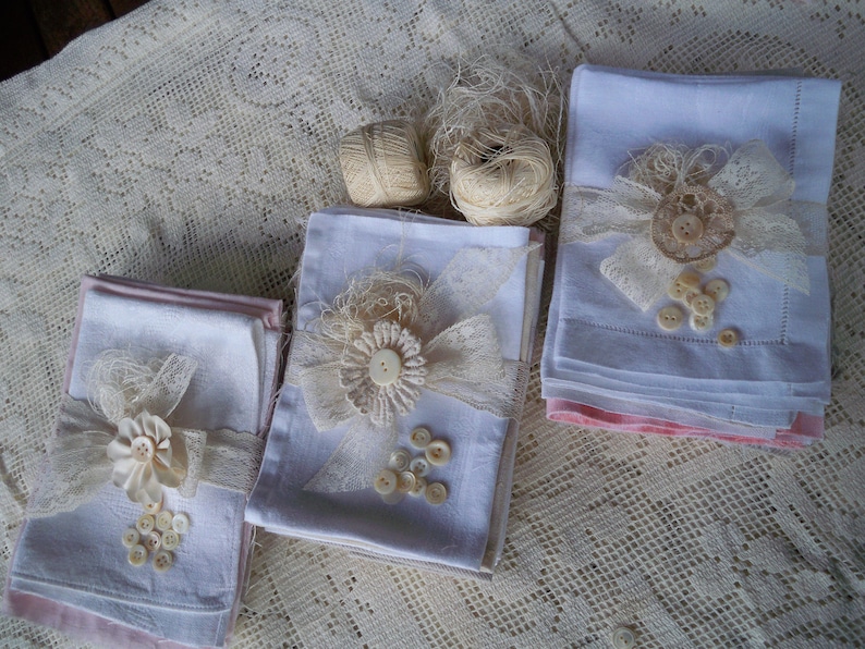 May include: Three stacks of white linen napkins, each adorned with lace, ribbon, and buttons. The napkins are decorated with floral accents and tied with lace. Two spools of twine are also visible, suggesting a craft or home decor theme.