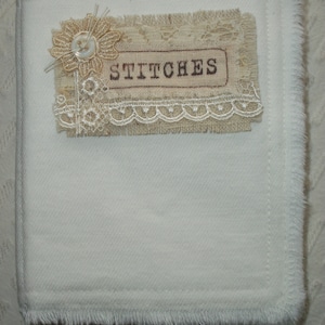 May include: A white fabric covered journal with a frayed edge. The front cover has a decorative label with a lace trim and the word "STITCHES" printed on it.