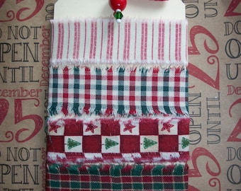 May include: A white tag with a red bow and a red bead. The tag has four strips of fabric attached to it. The fabric strips are red and white striped, red and green plaid, red and white checkered, and red with green Christmas trees. The tag is on a brown background with the words "Do Not Open Until December 25" printed on it.