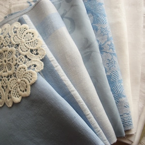 May include: A collection of folded fabric napkins in various shades of blue and white. One napkin features a cream-colored, circular lace applique. The fabrics include solid, striped, and patterned designs, creating a layered visual effect.