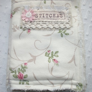 May include: A white fabric needle book with a floral pattern and a label that says "STITCHES". The label is decorated with lace and a pink button.