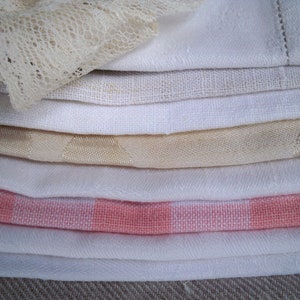 May include: A stack of folded white and cream-colored linens, including a lace-trimmed piece. Various textures are visible, such as linen, cotton, and a gingham pattern in pink and white. The fabrics are neatly arranged, suggesting a home decor or textile theme.