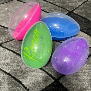 May include: Four clear plastic Easter eggs with different colored bases. The eggs are decorated with names, "Rocco", "Olivia", and two bunny silhouettes.