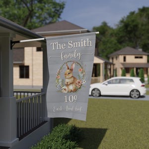 Op de afbeelding: Een grijze welkomstvlag met een bloemenkrans en een konijn. De tekst op de vlag luidt "The Smith family, 109 East Fruit Street."
