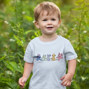 May include: A young child wearing a light blue t-shirt with a colorful graphic of dancing bears. The child is standing in a grassy area.