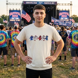 May include: A cream-colored short-sleeve t-shirt featuring a graphic of five dancing bears in red, white, and blue patriotic hats, each holding a small American flag. The design is centered on the chest of the shirt, which has a speckled texture.
