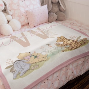 Pode incluir: Um cobertor de bebê rosa e branco com tema do Winnie the Pooh. O cobertor apresenta o Winnie the Pooh, o Porquinho e o Tigrão. O cobertor é personalizado com o nome "Olivia".
