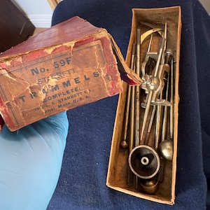 Puede incluir: Un juego de gramiles Starrett No. 59F vintage en una caja de cartón roja y beige desgastada. El set incluye varias varillas metálicas, trazadores puntiagudos y accesorios circulares. La tapa tiene el texto No. 59F STARRETT TRAMMELS COMPLETE THE L.S. STARRETT C., Athol, Mass., U.S.A.