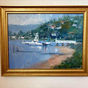 Puede incluir: Una pintura al óleo en un marco dorado que representa una escena costera con dos barcos amarrados cerca de un muelle blanco. El paisaje presenta una orilla arenosa, agua azul tranquila y una colina cubierta de árboles verdes. Firmado C.M. GRAWIN.