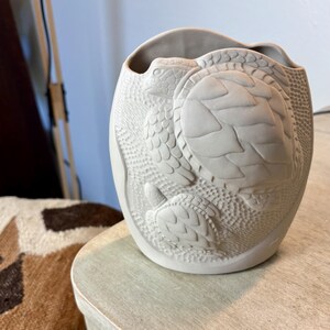 May include: A matte light gray ceramic vase featuring a raised relief design of two sea turtles swimming. The surface texture surrounding the turtles consists of small, dense dots, while the turtles themselves display detailed shell patterns.
