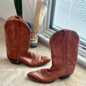 Puede incluir: Un par de botas vaqueras de media caña confeccionadas en cuero liso de color tostado. Cada bota presenta una punta afilada, un tacón apilado y patrones de costuras blancas decorativas en la caña. El cuero tiene un acabado natural con costuras.