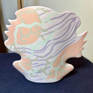 May include: A ceramic vase with an irregular, organic shape featuring a matte finish. The surface is decorated with a pattern of soft peach-colored patches, light green vein-like lines, and wavy lavender stripes across a pale off-white background.