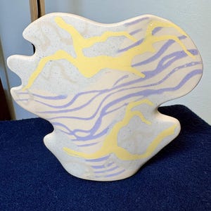 May include: A ceramic vase with an irregular, organic shape and a speckled off-white base. The surface features abstract, flowing patterns in light purple and bright yellow, creating a distinct contrast against the textured, matte-finish clay body.