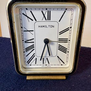 May include: A rectangular Hamilton table clock with a white face, black Roman numerals, and black hands. The clock features a metallic silver and black frame with a gold-toned base. The text HAMILTON is printed on the face, with MADE IN GERMANY below.