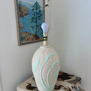 May include: A table lamp with an oval-shaped, textured beige ceramic base featuring raised, light green and soft pink floral motifs. The lamp has a metal harp, a white finial, and a single exposed light bulb extending from the top of the base.