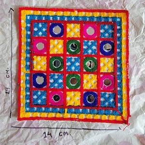 May include: A square, embroidered textile with a grid pattern. The design features red, yellow, blue, and green squares, each with a small mirror. The textile measures 5.5 inches by 5.5 inches, with an overall size of 7.5 inches by 5.5 inches.