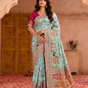 May include: A light blue saree featuring a vibrant floral print and intricate multi-coloured patterns along the border. The garment is paired with a magenta blouse that has short sleeves and a detailed embroidered trim along the sleeve edges.