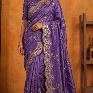 May include: A deep purple saree featuring a textured fabric finish, adorned with intricate gold embroidery and small circular mirror accents along the scalloped borders and across the body, paired with a matching short-sleeved blouse.