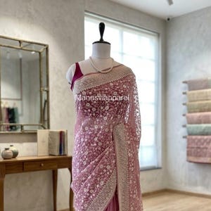 May include: A dusty rose saree with intricate white floral embroidery and a delicate lace border. The saree is draped on a mannequin, showcasing its flowing fabric and detailed design. The text "Manasviapparel" is visible on the saree.