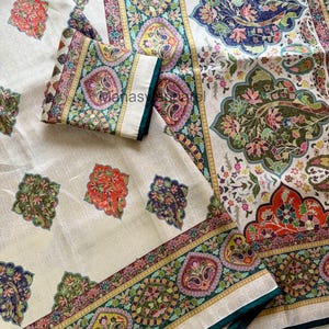 May include: Cream-colored textile with intricate floral and paisley patterns in shades of red, green, blue, and purple. The design includes a border with a dark green trim. The textile has the text "Manas Apparel".