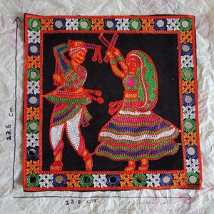 May include: A square, embroidered textile featuring two figures in traditional dance poses against a black background. The figures are rendered in vibrant orange, red, green, and white thread. The border is decorated with mirrors and geometric patterns. The textile measures 23.5 cm x 23.5 cm.
