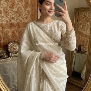 Puede incluir: Traje tradicional indio de color crema con bordados dorados. El conjunto incluye una parte superior de manga larga y tela drapeada, con ribetes dorados. El texto "Manasviapparel" es visible.