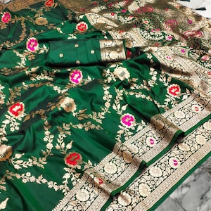 May include: A green silk saree with gold and silver embroidery. The saree has a floral pattern and a border with a geometric design. The saree is draped over a white surface.