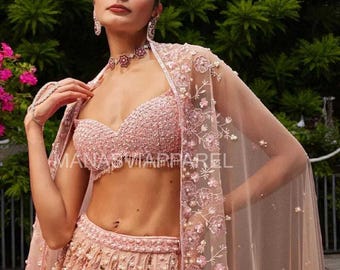 May include: A light pink, embellished bridal ensemble. The set includes a beaded crop top, a full skirt with floral embroidery, and a sheer cape with floral accents. The text "MANAS APPAREL" is visible. The outfit is accessorized with matching jewelry.