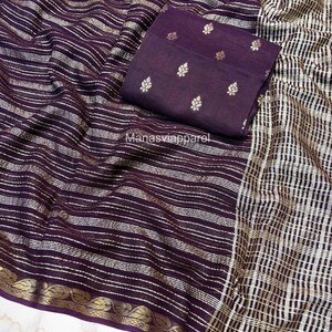 Könnte beinhalten: Ein dunkelvioletter Sari mit horizontalen silbernen Streifen und einem goldenen Rand. Ein passender dunkelvioletter Stoff mit silbernen Blattmustern ist oben gefaltet. Der Text "Manasviapparel" ist sichtbar.