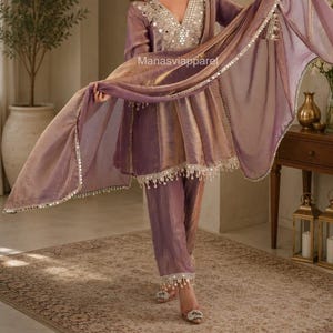 Peut inclure: Un ensemble trois pièces mauve composé d'une tunique à col en V ornée de miroirs argentés, d'un pantalon assorti et d'une dupatta transparente. Les bords de la tunique et de la dupatta sont décorés de miroirs et de pompons. Texte : Manasviapparel.