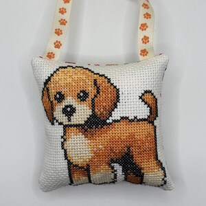 May include: A small square fabric pillow featuring a cross-stitch design of a brown and white puppy. The pillow has a cream-colored ribbon handle attached to the top, decorated with a repeating pattern of small orange paw prints.