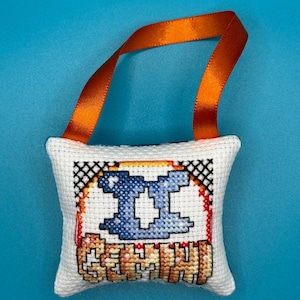 May include: A white, cross-stitched pillow with the Gemini symbol in blue and the word "GEMINI" in orange. An orange ribbon is attached for hanging. The pillow is on a blue background.