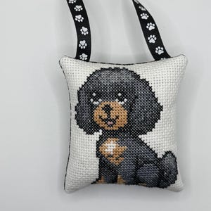 May include: A square cross-stitch ornament featuring a black and tan puppy on a white fabric background. The ornament is finished with a black ribbon hanger decorated with a repeating white paw print pattern, allowing it to be hung as a decoration.