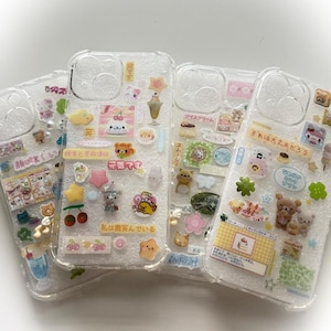 May include: Four clear phone cases decorated with colorful, kawaii-style stickers. The stickers include food items, stars, and cartoon characters. Japanese text is visible on some of the stickers. The cases are arranged on a white surface.