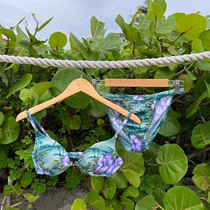 May include: A green and purple floral print bikini top and bottom hanging on a wooden hanger. The bikini is on a white rope against a backdrop of green foliage.