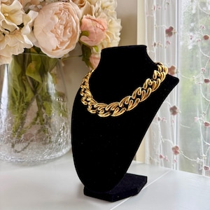 May include: A gold-toned chunky chain necklace featuring thick, interlocking oval links with a polished, reflective surface. The necklace is displayed on a black velvet-covered bust, showcasing the uniform size and shape of the metal links.