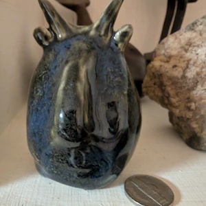 May include: A glazed ceramic sculpture in dark blue and black. The artwork features a rounded form with two upward horns and two small ear-like projections. A silver coin is placed close by.