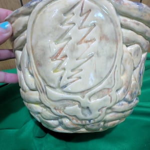 May include: A beige ceramic bowl with a glossy finish featuring a raised, embossed lightning bolt design within a circular frame. The surface has subtle brown and orange mottled color variations throughout the textured, sculpted clay exterior.