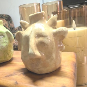 May include: A ceramic sculpture of a face with exaggerated features. The sculpture is beige with a textured surface and has a whimsical, almost cartoonish appearance.