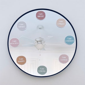 May include: A round mirror with a black frame reflects a ceiling fan and several colourful stickers with positive affirmations. The stickers read "I am Beautiful", "I am Smart", "I am Happy", "I am Brave", "I am Strong", "I am Unique", "I am Confident", and "I am Loved".
