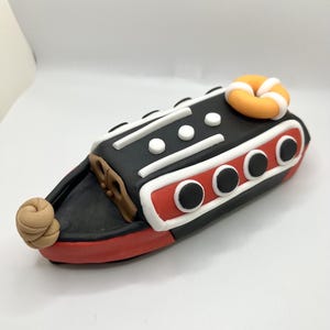 May include: A black and red toy boat with white accents and a brown rope. The boat has three round portholes on the side and a life preserver on the top.