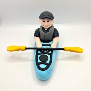 May include: A blue and black kayak with a person in a grey life jacket and black hat paddling with a yellow paddle.