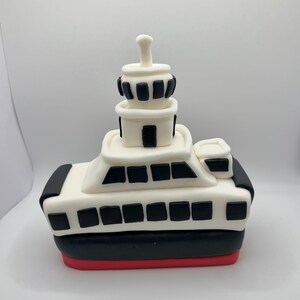 May include: A white and black fondant cake decorated to look like a ferry boat. The cake has a red bottom, black sides, and a white top with black windows. The cake has a small white tower on top with black windows.