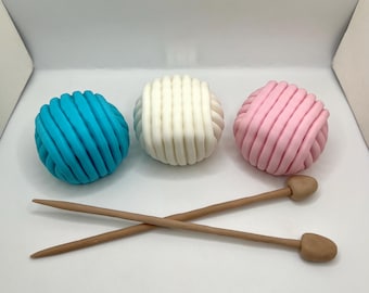 Könnte beinhalten: Drei Wollknäuel, eines blau, eines weiß und eines rosa, mit zwei Stricknadeln aus Holz.